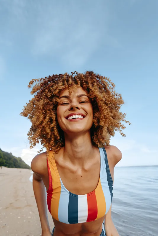 Smiling woman with curly hair at beach near ocean under bright sunlight Concept of vacation lifestyle and freedom Relaxed mood with clear blue sky close up face in striped swimwear