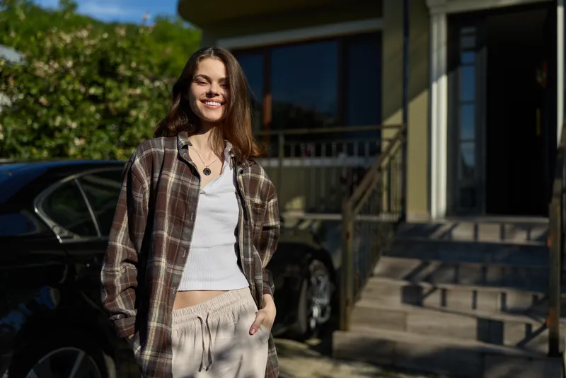 A smiling woman in a plaid shirt and white top poses outdoors, showcasing a casual, relaxed lifestyle in daytime sunlight showcasing confidence and everyday style