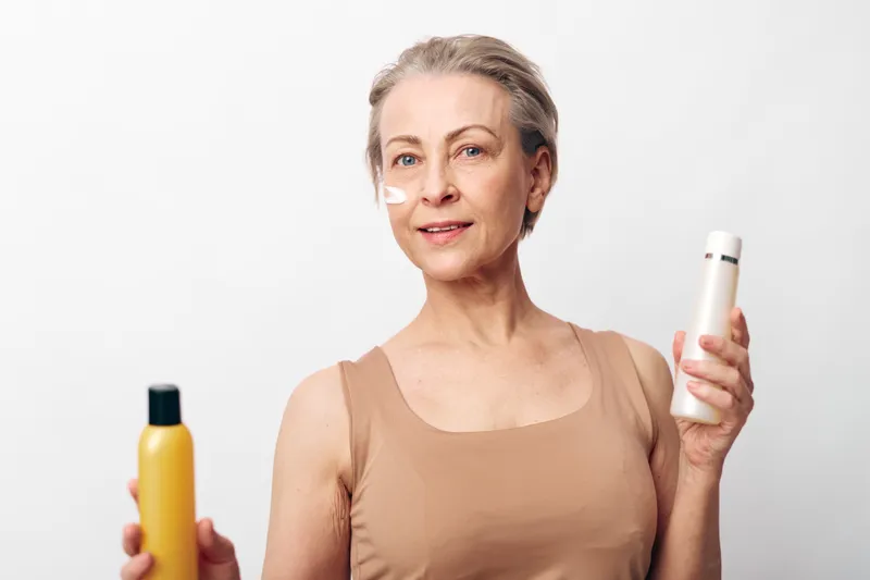 Mature woman with short grey hair holds two cosmetic bottles and applies cream on cheek. Bright studio background highlights skincare, beauty routine, self-care, and natural aging concepts.