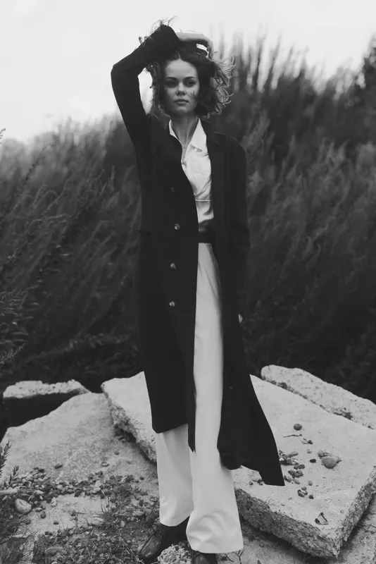 Woman in long coat and white dress stands outdoors, wind in her hair, monochrome fashion scene, moody and stylish, natural setting with rocks and grass, bold pose and cinematic atmosphere