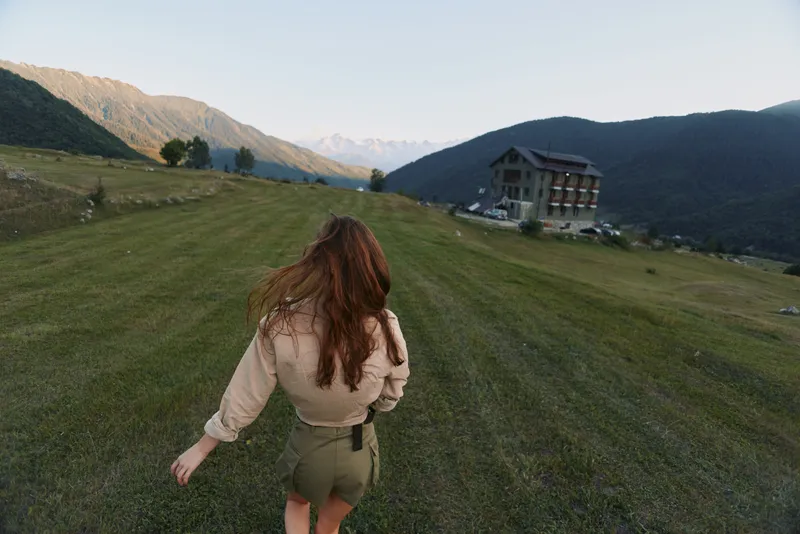 Woman walks away across a green field toward distant mountains, capturing a casual outdoor lifestyle moment in a rural landscape, with expansive sky and quiet atmosphere.