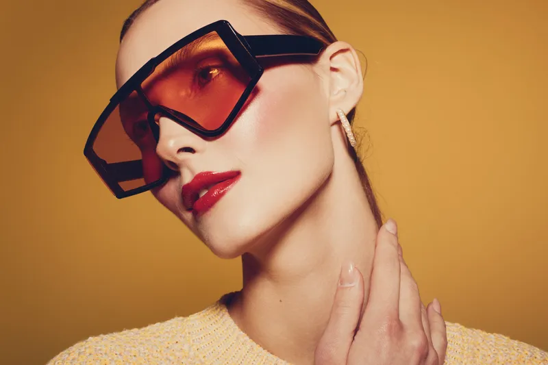 Stylish woman with oversized orange sunglasses and red lipstick poses against yellow background. Fashion portrait with modern accessories and glossy makeup highlights elegance and confidence.