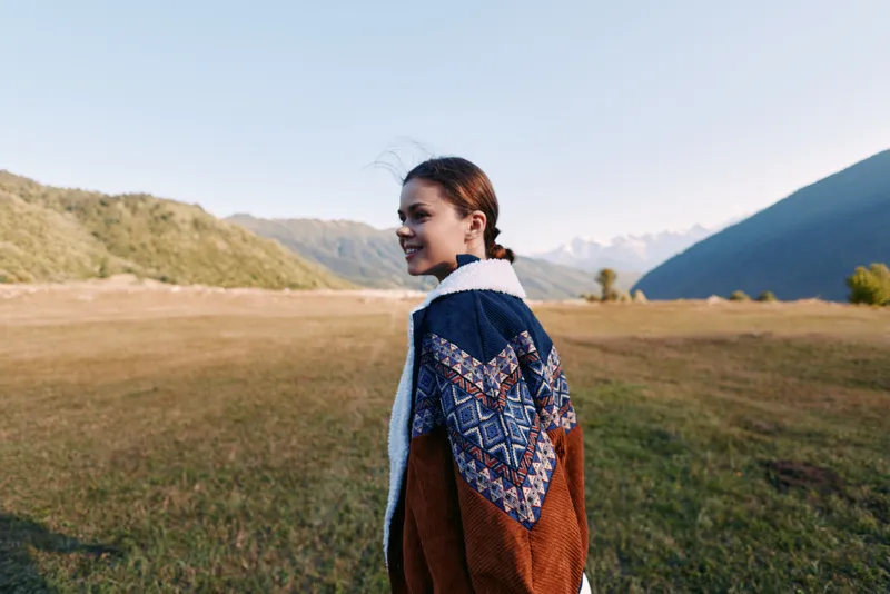 Woman smile portrait in patterned jacket walking across a grassy field with mountains in the background. Outdoors lifestyle, scenic landscape, casual fashion and happy travel moment.