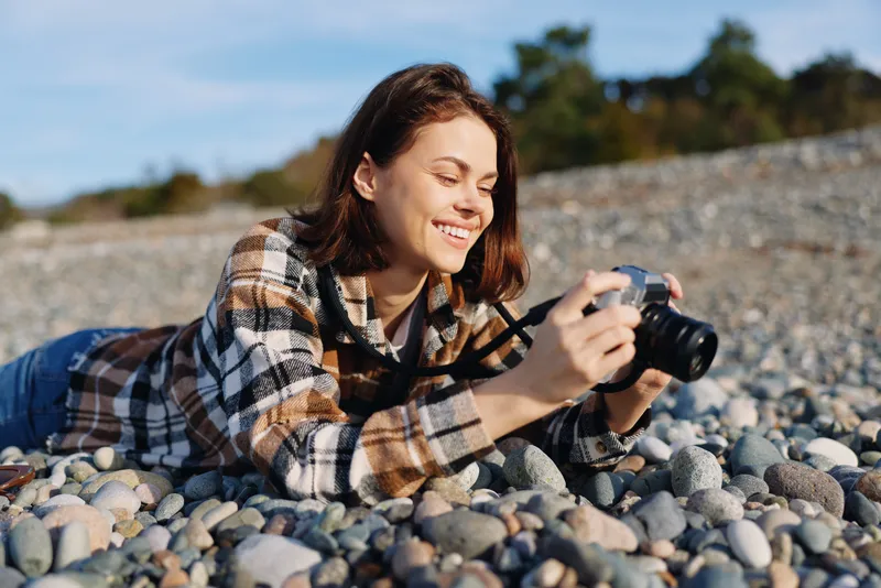 Photographer lying on a pebble beach with a camera, capturing outdoor moments in natural light. Casual plaid jacket, relaxed smile, seaside scenery, creative hobby and travel vibe.
