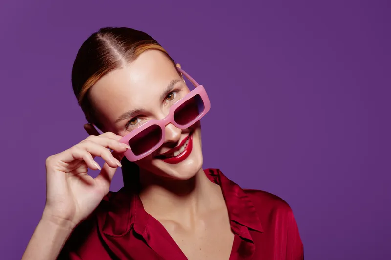 Smiling woman with slicked hair wearing pink sunglasses and red shirt poses against purple background. Concept of fashion, style, summer, and confident female portrait with bright colors.