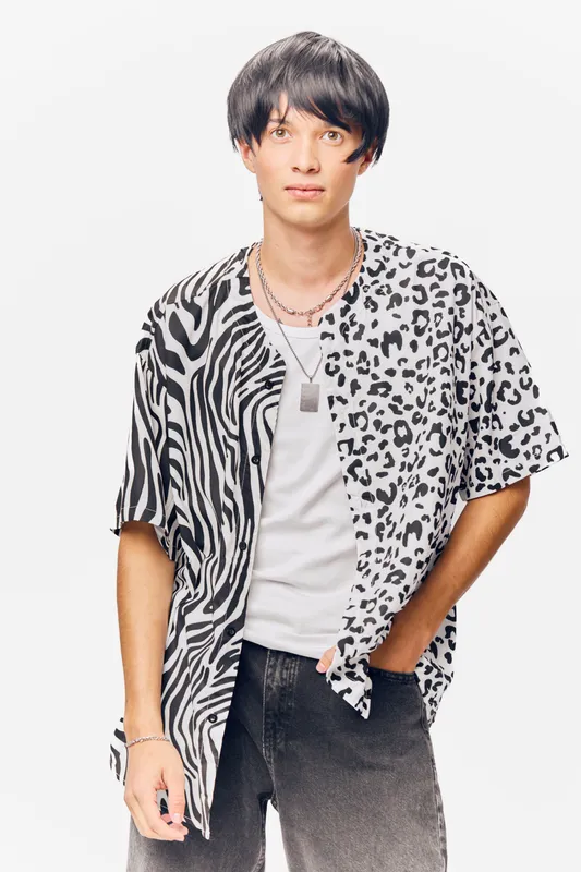 young man wearing a black and white animal print shirt with a necklace and casual jeans, standing with a hand in pocket on a white background, modern style
