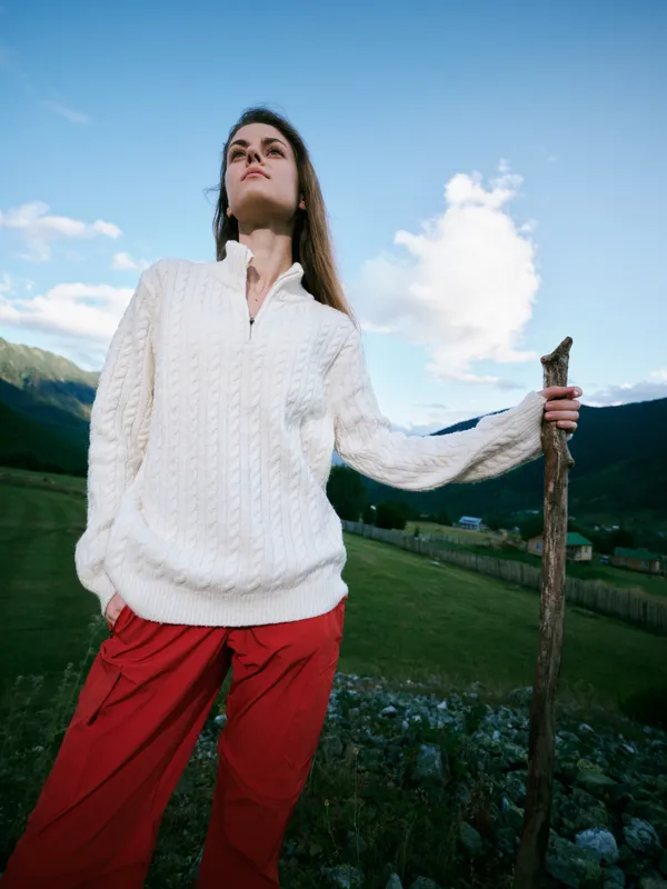 Woman in white knit sweater and red pants stands with a wooden staff in a green valley, gazing at the bright sky with distant mountains, outdoors, exploration and freedom