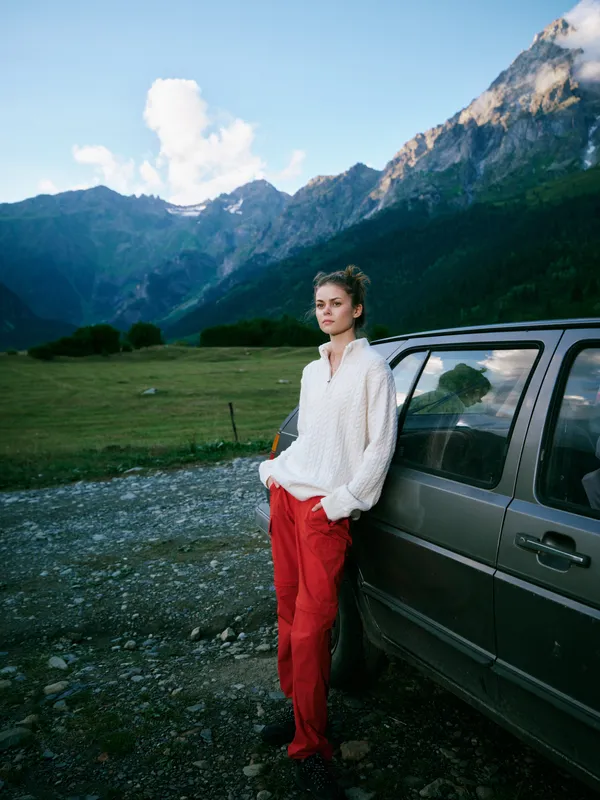 Woman leaning against a car in a mountain valley, casual fashion with white sweater and red pants, open space, dramatic scenery and fresh air