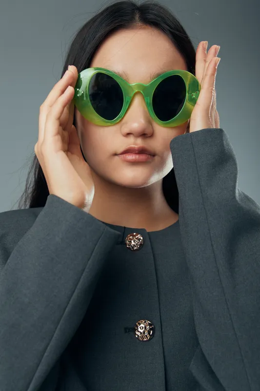 Woman wearing large green sunglasses and dark gray coat posing against neutral background with serious expression and long black hair for fashion portrait.