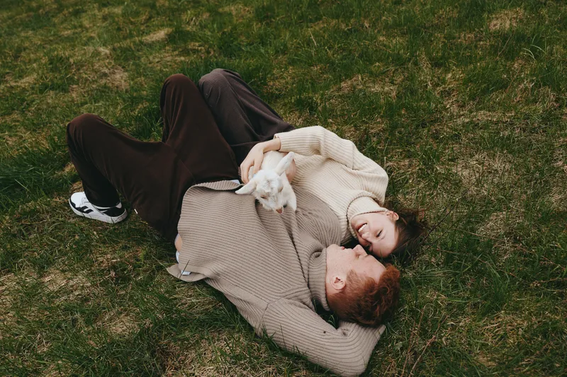 A relaxed couple lies on grass with a small goat kid resting between them, creating a cozy lifestyle moment outdoors, blending affection, nature, and gentle companionship.