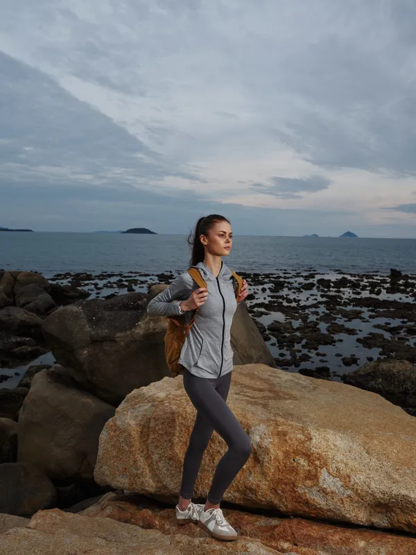 Athletic woman with a backpack enjoying nature by the seaside, wearing gray sportswear and showing a contemplative emotion against a dramatic coastal backdrop