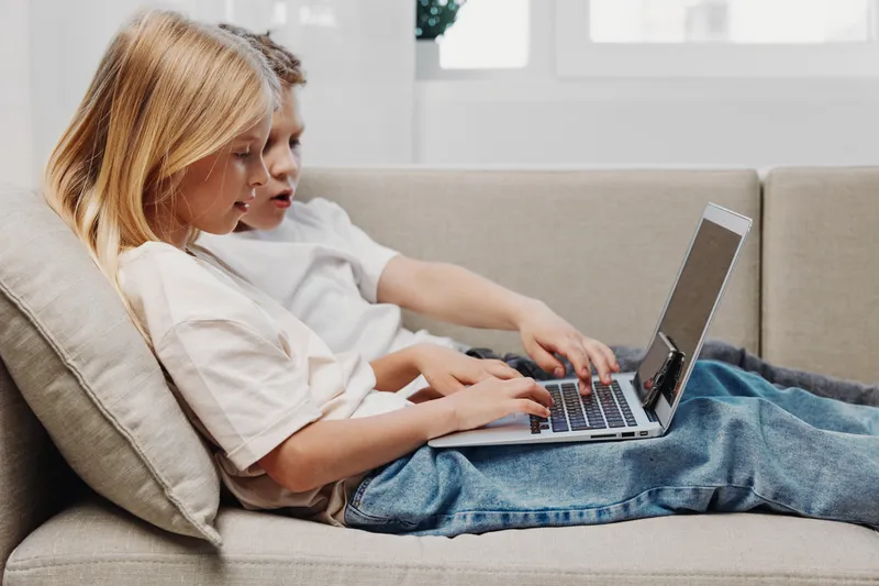Two focused children sitting on sofa use laptop together in bright room, learning and exploring technology concepts with concentration and teamwork.