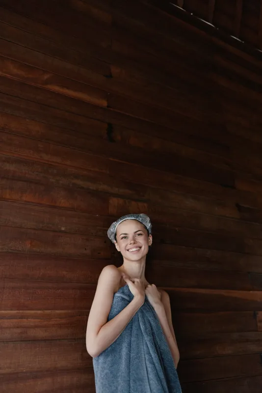 smiling woman wrapped in blue towel and shower cap stands against warm wooden wall, showing relaxation and freshness after bath, natural light highlights happy expression