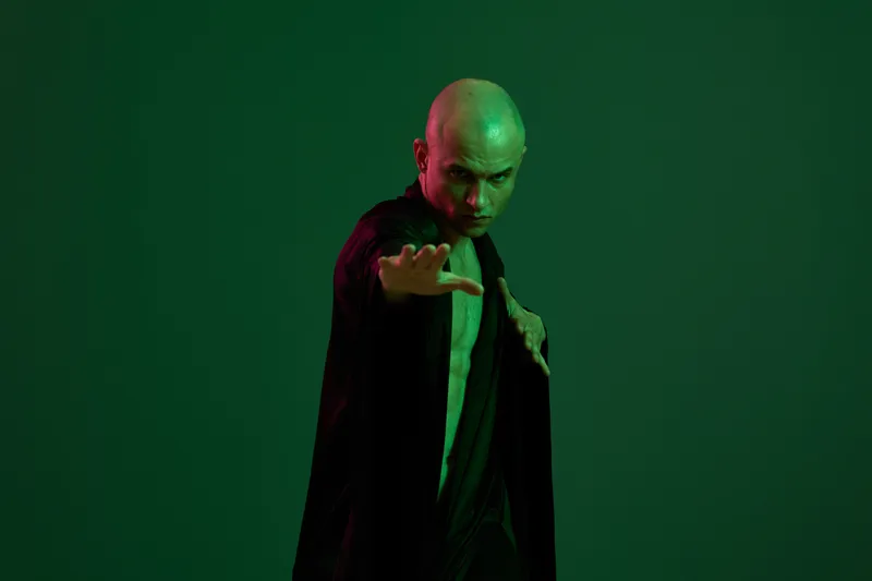 Striking man performing a dramatic pose with an intense expression, dressed in dark clothing against a green background, showcasing power and elegance.
