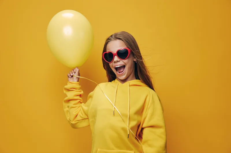 Girl in yellow hoodie and red heart sunglasses holding yellow balloon with cheerful expression on bright yellow background for celebration and fun mood.