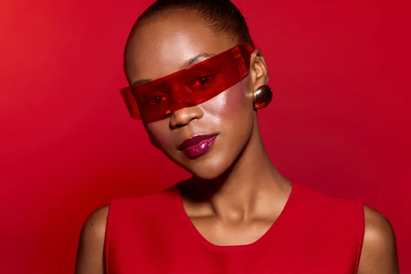 Portrait of a confident black woman with dark skin wearing red translucent sunglasses and a sleeveless red top against a red background. Concept of fashion, style, and modern beauty.