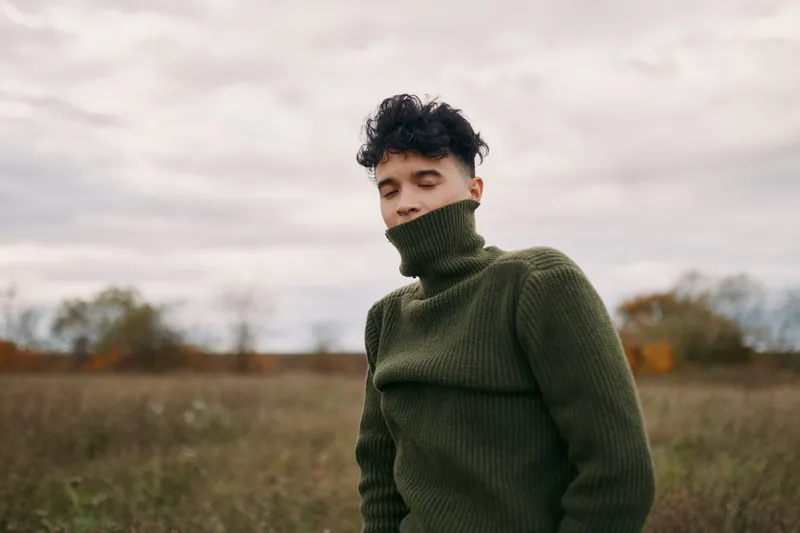 A young man in a green turtleneck sweater stands in an open field with eyes closed, posed naturally to convey authentic feeling, genuine emotion and a credible, understated realism.