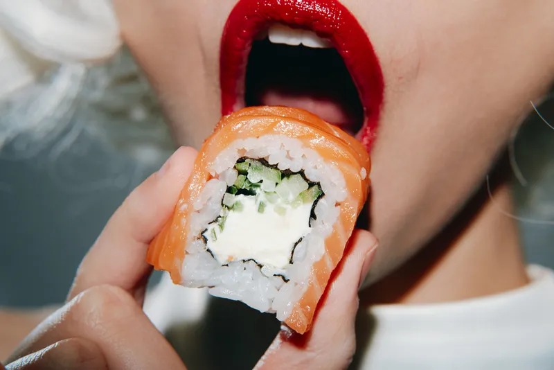 Модная модель с яркой красной помадой откусывает вкусный ролл с суши с лососем и огурцом, идеально подходит для любителей еды