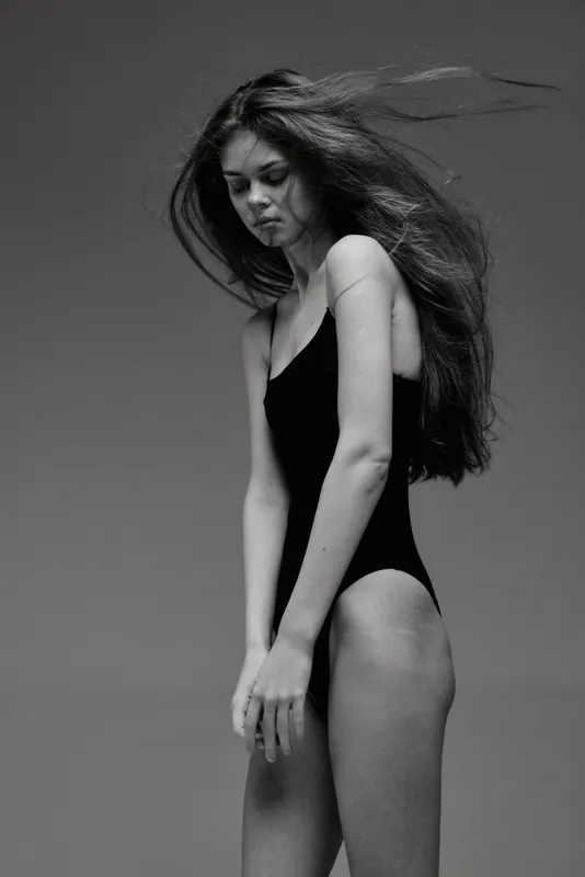 Black and white portrait of a young woman with long hair wearing a black swimsuit looking down thoughtfully.
