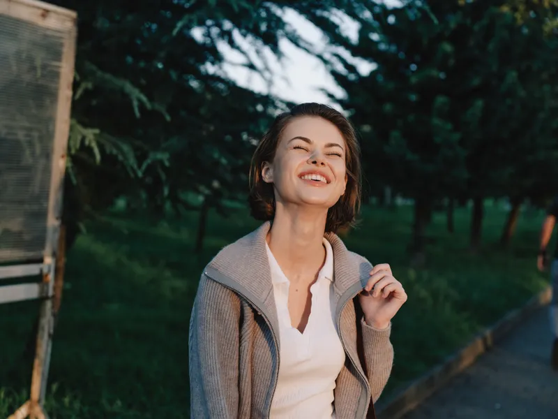 A cheerful woman stands outdoors in a park, smiling warmly as she enjoys a bright, sunny day. She wears a light jacket over a white blouse, exuding upbeat confidence and happiness.