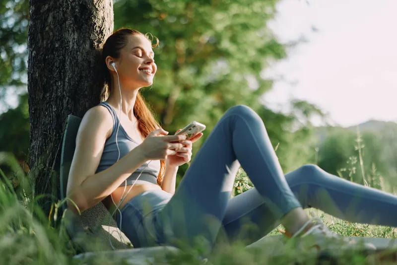 Young woman with earphones enjoying music outdoors, wearing sportswear, relaxed and happy, surrounded by a green summer landscape