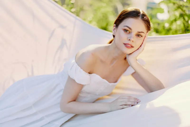 Young woman in a delicate white dress posing gracefully outdoors, showcasing a serene expression and soft natural light, perfect for spring or summer themes