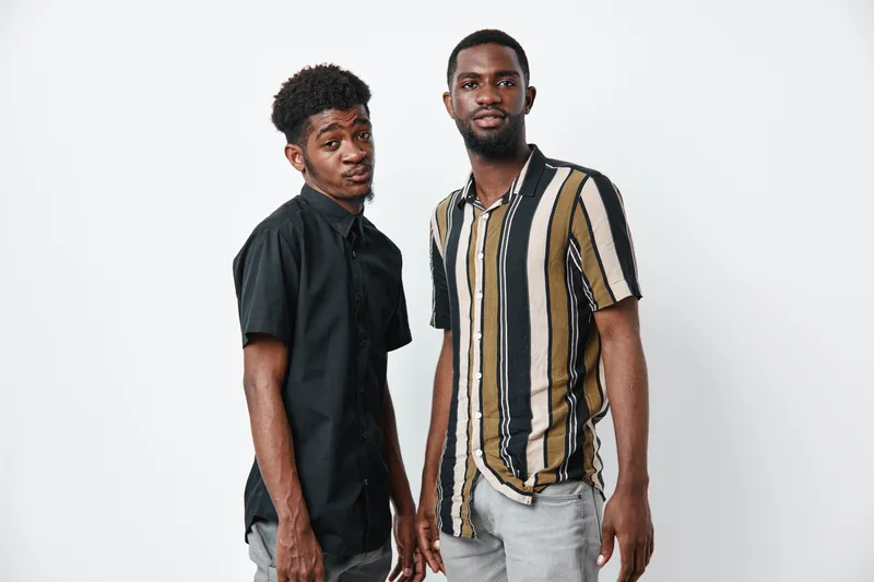 Two young black men with dark skin standing together in casual shirts against white background. African ethnicity, friendship, confidence, and modern style in studio portrait.