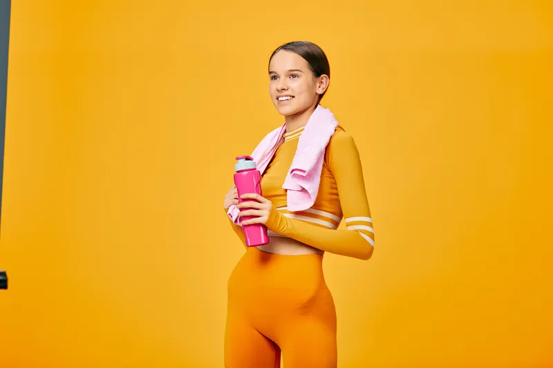 Energetic woman in bright orange workout set holding a pink water bottle and towel after training against a vivid orange backdrop, showcasing fitness lifestyle and athletic confidence.