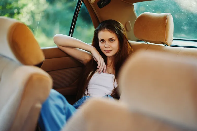 Young woman relaxing in a vintage car with natural sunlight, wearing casual clothes and a joyful expression, surrounded by warm colors of summer.