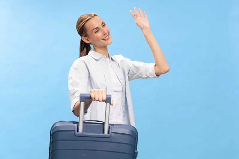 Young happy woman with suitcase waving hand on blue background. Travel concept with casual clothes, positive mood, and summer vacation preparation, isolated studio portrait.