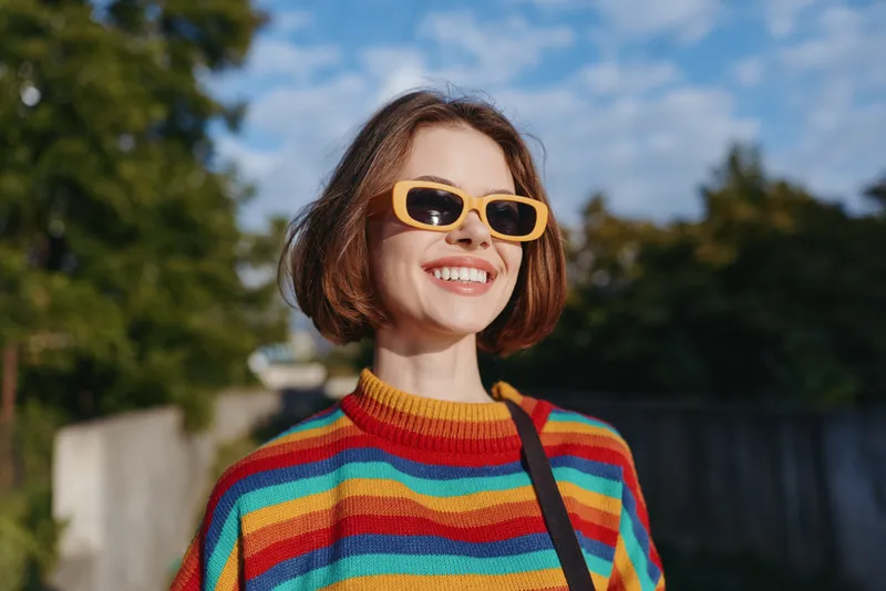 woman smiling in her 20s with short hair and bob haircut, wearing a colorful striped sweater and yellow sunglasses, walking outdoor in sunny urban park, casual outfit lifestyle portrait.
