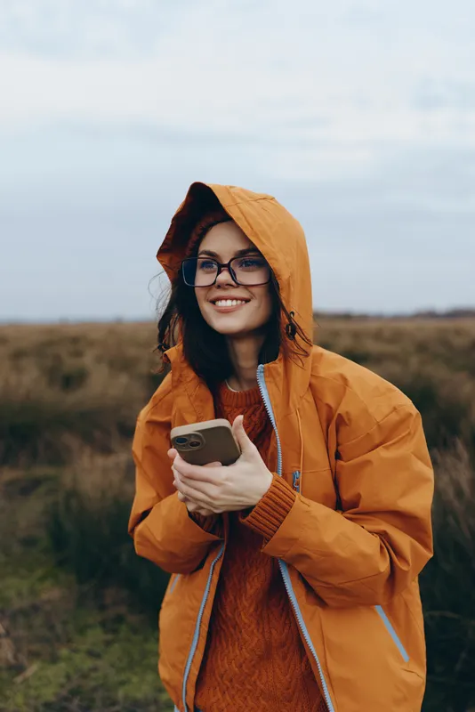 woman lifestyle outdoors raincoat hoodie smartphone smile glasses field cloudy, smiling woman holds smartphone, orange hooded jacket, countryside meadow, overcast sky, daylight, relaxed travel mood