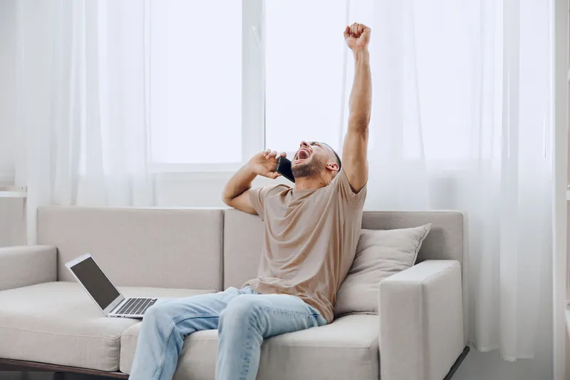 Excited man celebrating a successful phone call while relaxing on a couch, embodying the balance of personal and professional life in a soft masculine environment.