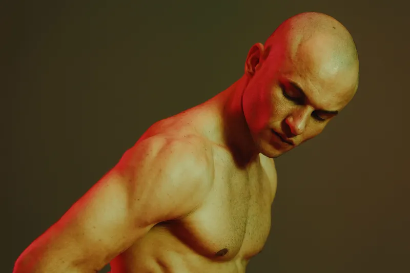 Seductive man posing with intense expression under dramatic light, showcasing strong physique and artistic shadows. A captivating visual story in a vibrant palette.