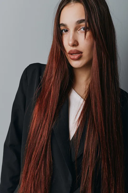 Young woman with long straight red hair wearing black jacket and white shirt posing against plain gray background for fashion portrait and beauty concept.