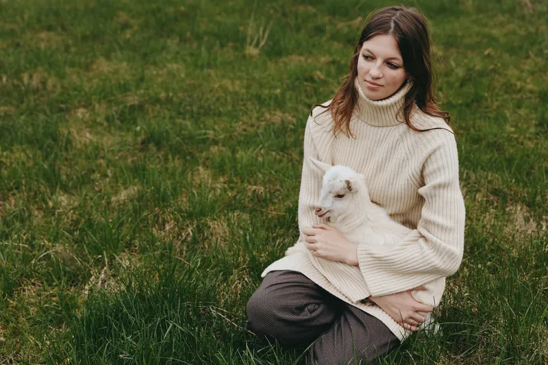 Calm woman sits on a grassy field, wearing a cozy beige sweater, holding a playful goat kid, sharing a gentle bond in a peaceful outdoor lifestyle moment.