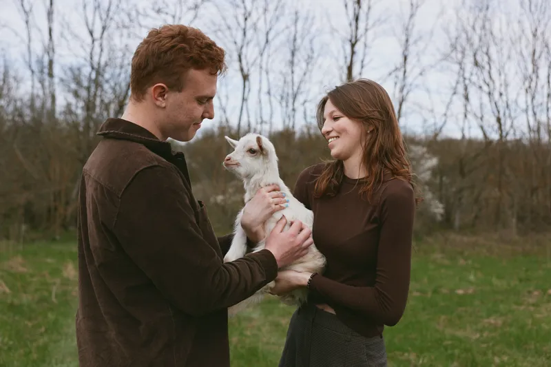 Playful couple outdoors cradle a small goat kid, smiling at each other, warm light, casual clothes, gentle breeze, rural setting, intimate lifestyle moment, natural colors, rustic background