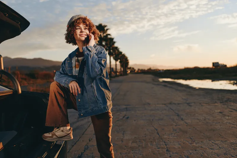 Woman lifestyle at sunset, smiling woman in denim jacket and jeans stands near car by roadside, outdoor travel mood, casual adventure on long road, warm evening light, golden