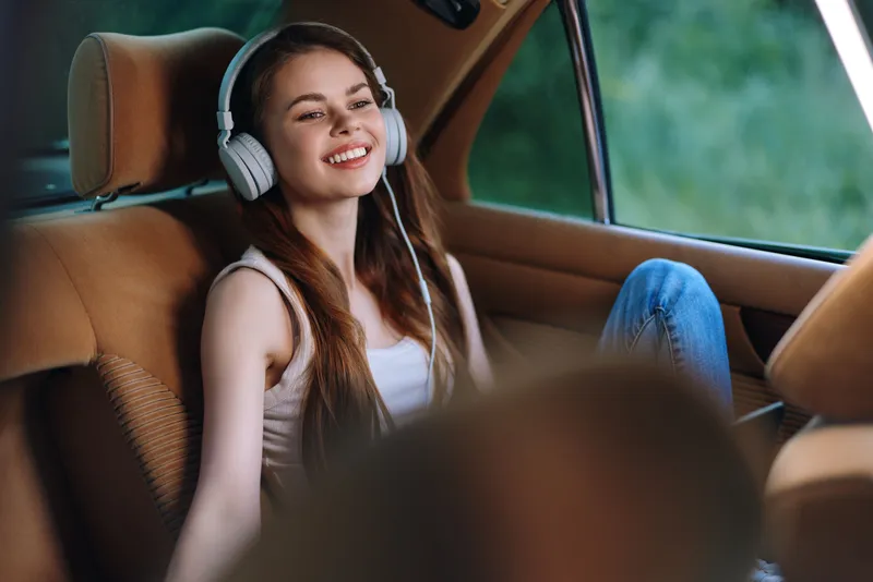 Young woman enjoying music in a car, with headphones on, smiling joyfully, featuring a natural backdrop and soft colors, perfect for lifestyle themes.