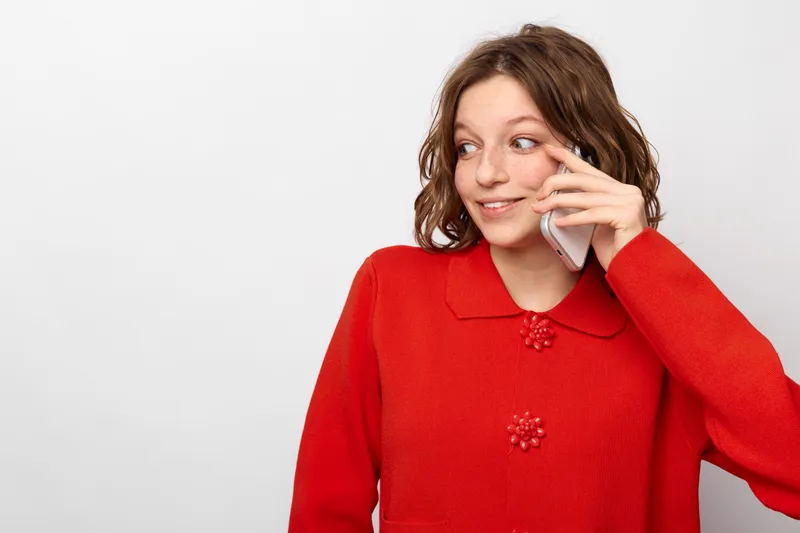 Young woman in red sweater talking on smartphone looks aside with smile on white background. Concept of communication, technology, casual style, and positive emotions in everyday life.