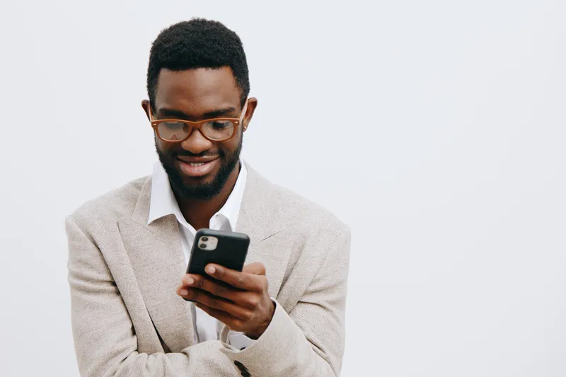 Smiling man with glasses using smartphone, casual beige sweater, isolated on plain white background. Studio portrait showcasing modern technology, communication, engagement, happiness, and technology 