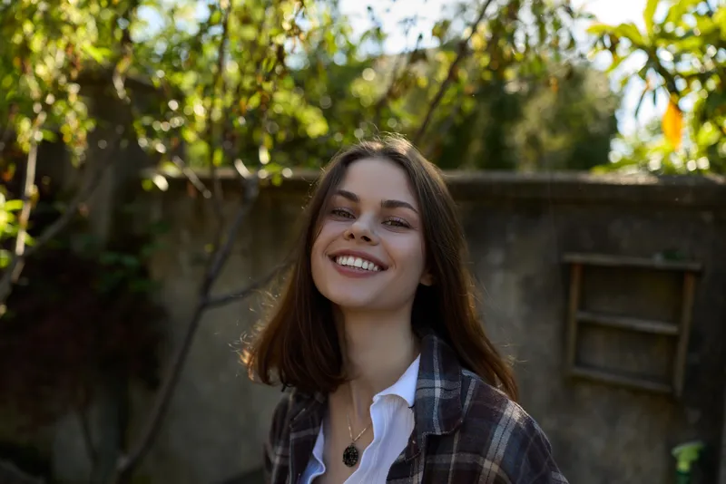 Happy young woman outdoors in a plaid shirt, smiling toward the camera, warm sunlight, casual lifestyle portrait in a leafy garden setting, carefree positive mood