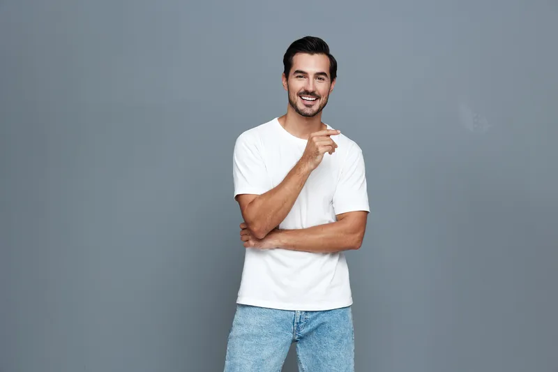 Smiling happy cheerful young man in a white t-shirt poses confidently against a plain gray background, showcasing a joyful expression. People lifestyle concept
