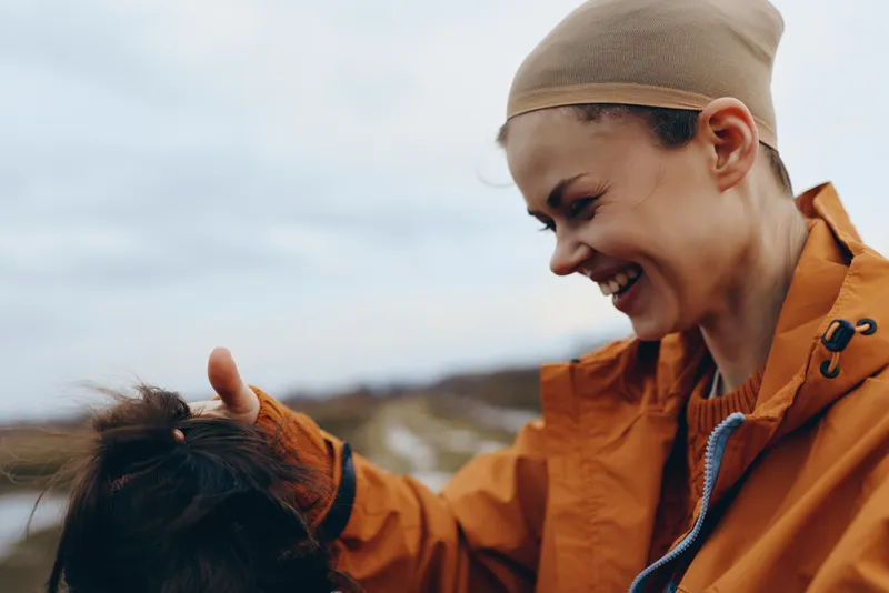 Smiling woman in orange jacket gently walking with a dog outdoors near open nature, close side view shows bond and friendship, bright morning light, lifestyle moment