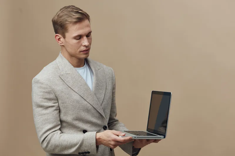 Professional young man in business casual clothing holding a laptop computer while standing against a plain beige background, focused on work and technology, in a studio shot. Business lifestyle conce