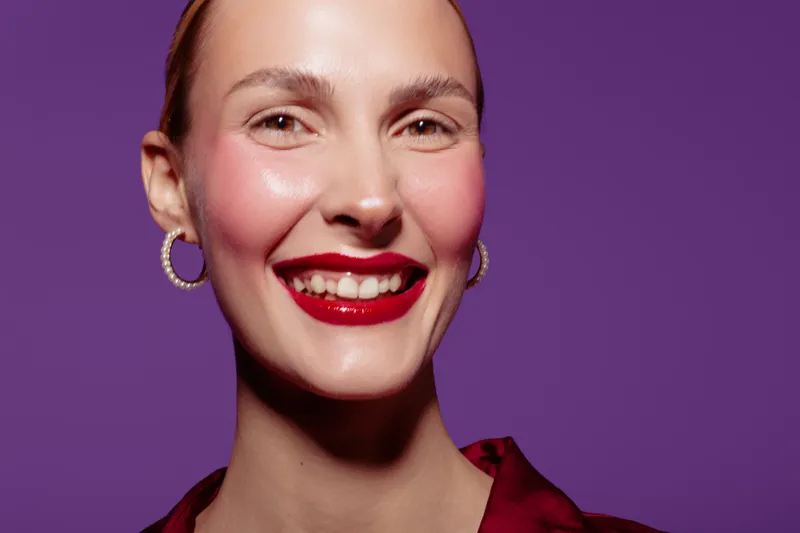 Smiling woman with red lipstick and hoop earrings poses against purple background. Close-up portrait of happy female model with glossy makeup and smooth skin. Concept of beauty and fashion.