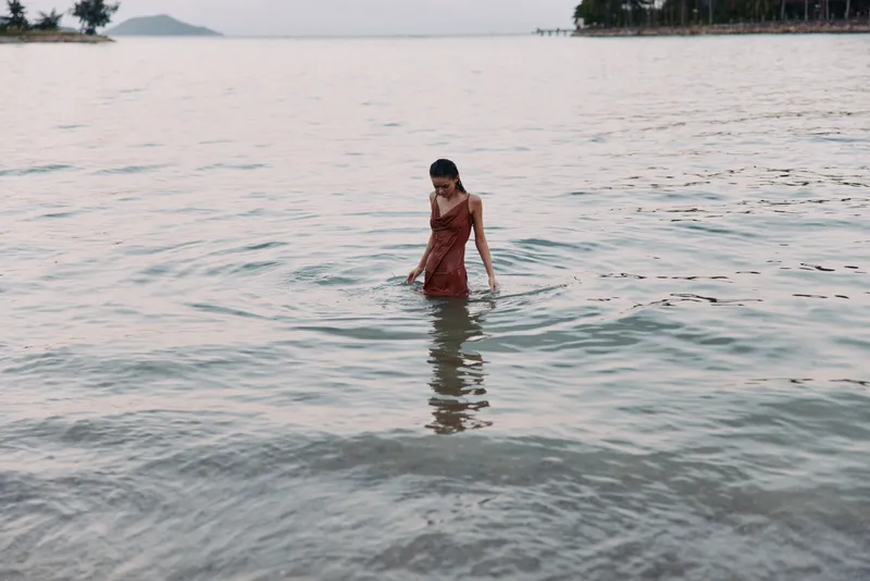 Woman stands waist deep in serene sea water, wearing a red dress. Soft light reflects on the rippling surface as distant shorelines fade into the horizon.