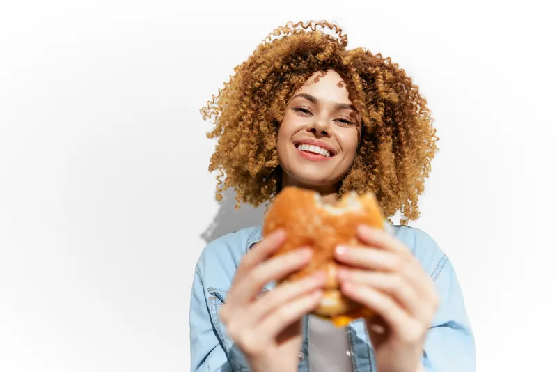 Счастливая женщина с вьющимися волосами наслаждается вкусным бургером, передавая эмоции восторга и волнения