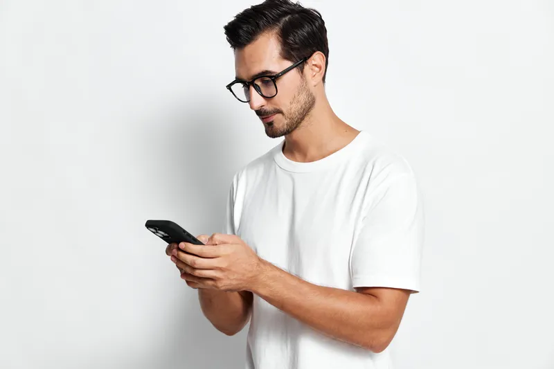 Thoughtful man in a casual white t-shirt wearing glasses, looking at his mobile phone with focused expression, isolated on plain solid background.