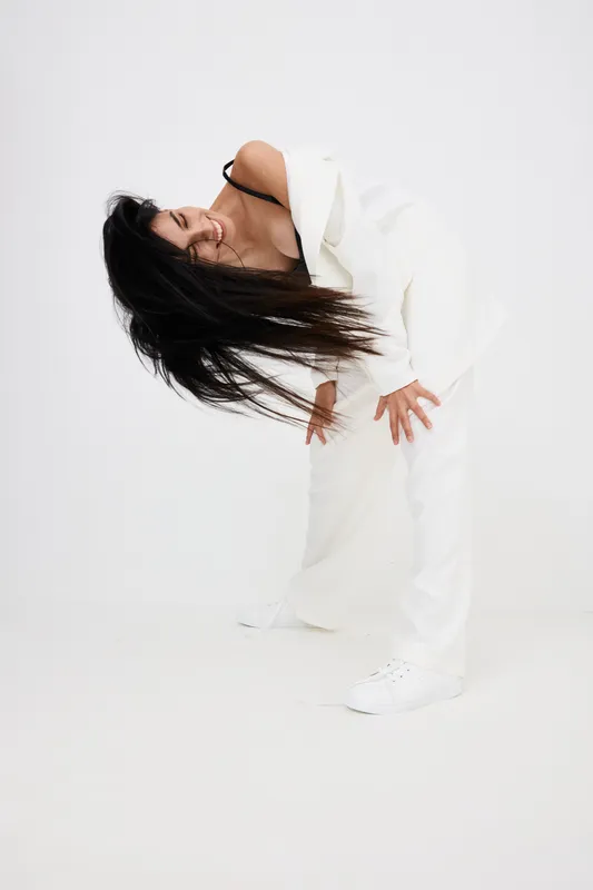 Woman bending forward in a stylish white suit with long black hair, posing on a white seamless background with dynamic movement and casual white sneakers.